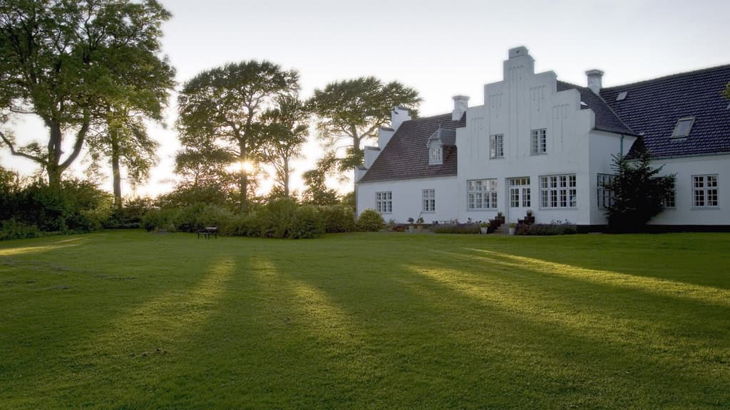 Brandbjerg Manor's facade with light falling through the trees on a beautifully freshly mowed green lawn.
