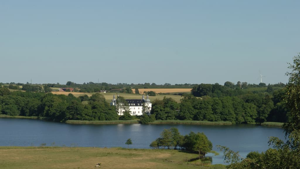 Engelsholm Castle from a distance with a view of the lake, trees, and surrounding fields.